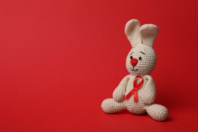 Cute knitted toy bunny with ribbon on red background, space for text. AIDS disease awareness