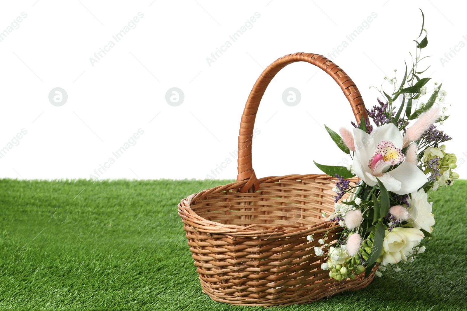 Photo of Wicker basket decorated with beautiful flowers on green grass against white background, space for text. Easter item