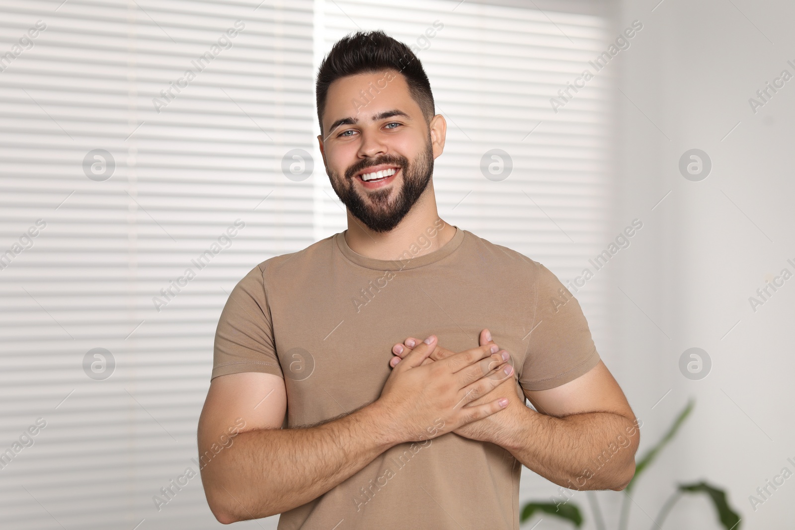 Photo of Thank you gesture. Happy grateful man holding hands near heart indoors