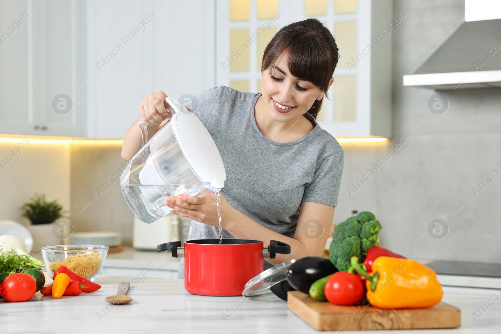 Photo of Happy young housewife pouring water in pot at white marble table in kitchen. Cooking process