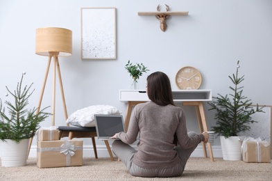 Photo of Woman with laptop sitting on floor in room decorated with potted fir trees, back view