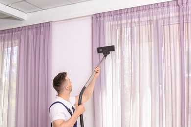 Photo of Male worker removing dust from curtains with professional vacuum cleaner indoors