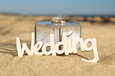 Photo of Box with decor, gold rings and word Wedding on sandy beach, closeup