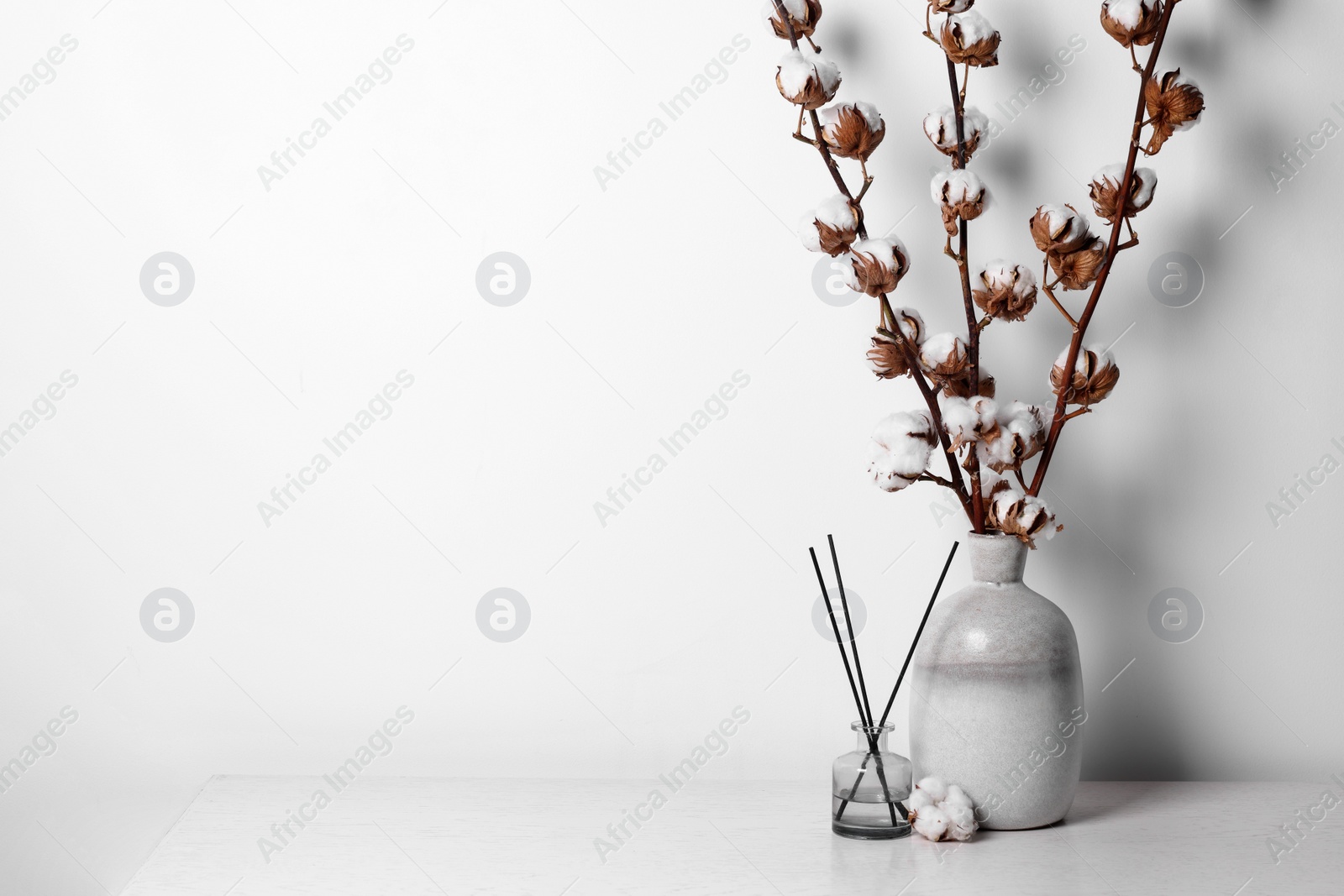 Photo of Reed diffuser and vase with cotton branches on table against white background. Space for text
