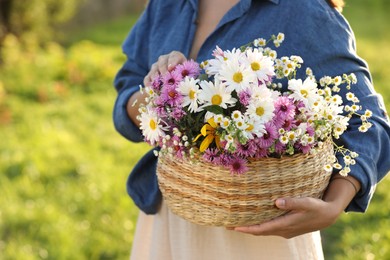 Photo of Woman holding wicker basket with beautiful wild flowers outdoors, closeup