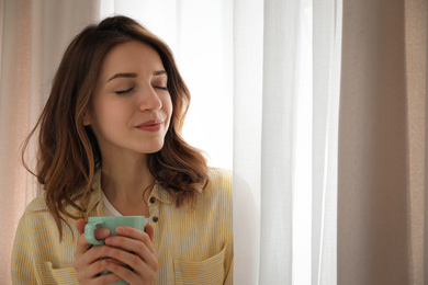 Woman holding cup near window with beautiful curtains at home