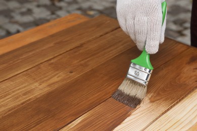 Man applying wood stain onto wooden surface against blurred background, closeup. Space for text