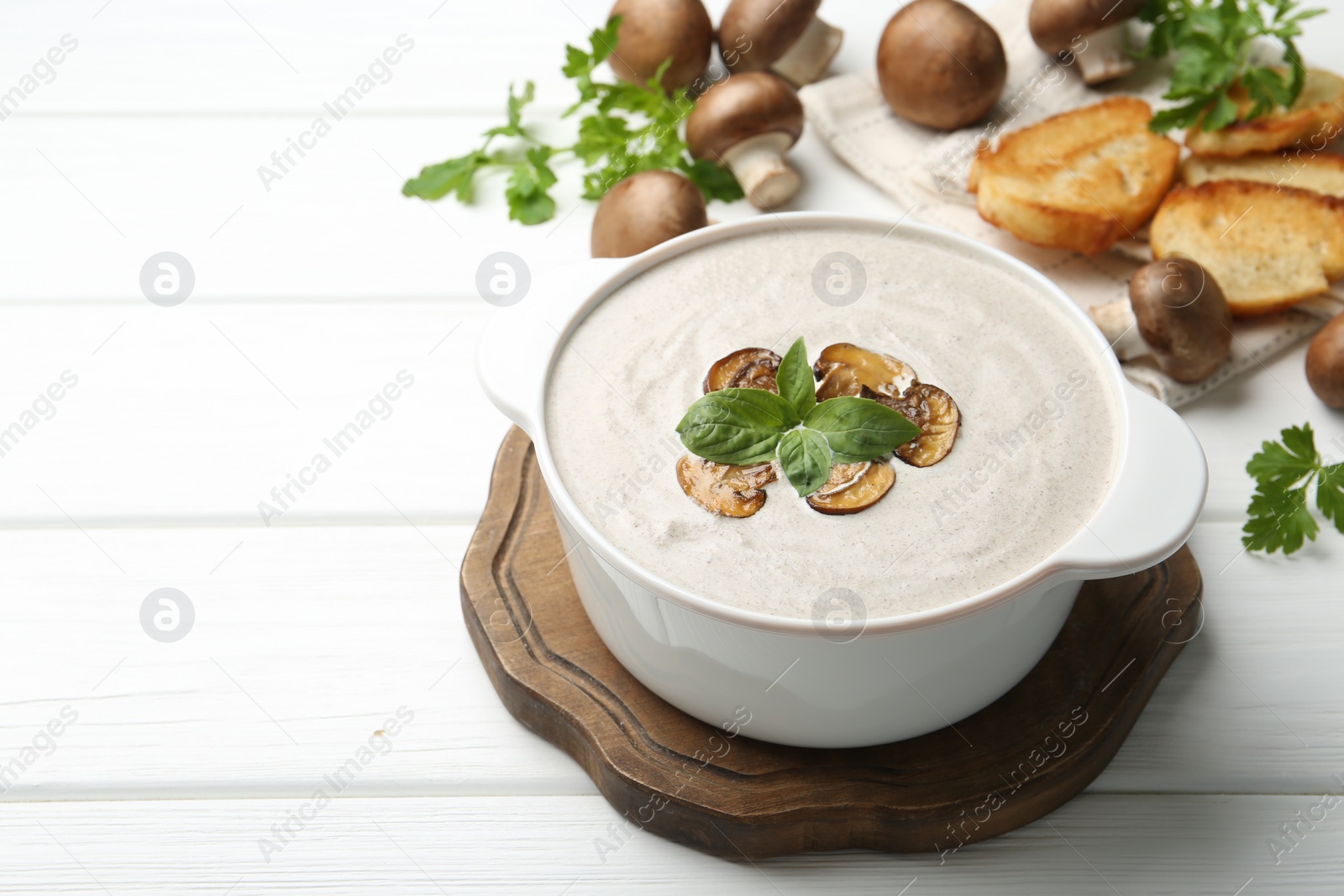 Photo of Delicious homemade mushroom soup in ceramic pot, croutons, fresh champignons and parsley on white wooden table. Space for text