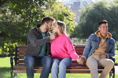 Photo of Woman reaching for kiss with another man while holding boyfriend's hand on bench in park. Love triangle