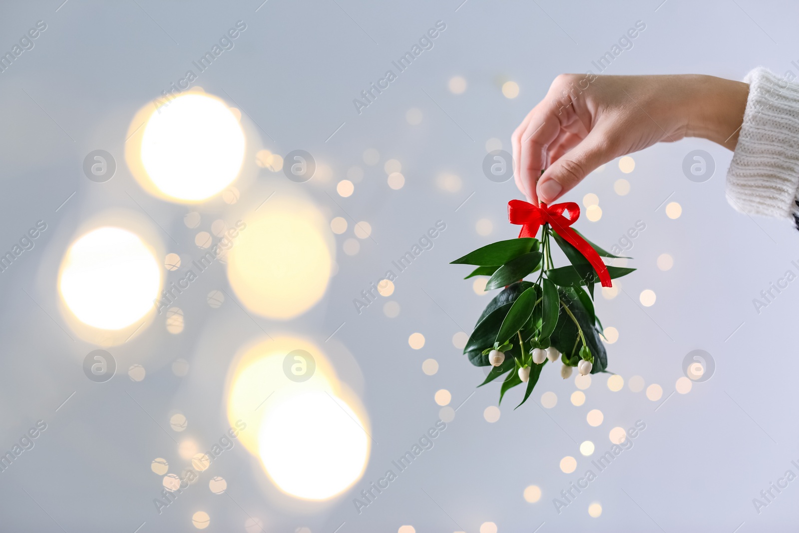 Photo of Woman holding mistletoe bunch with red bow against blurred festive lights, closeup and space for text. Traditional Christmas decor