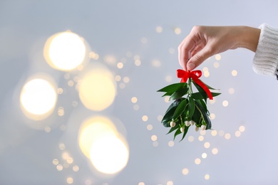 Photo of Woman holding mistletoe bunch with red bow against blurred festive lights, closeup and space for text. Traditional Christmas decor