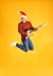 Photo of Man in Santa hat jumping with electric guitar on yellow background. Christmas music