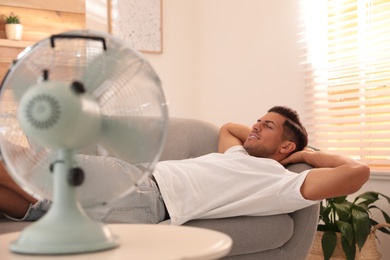 Photo of Man enjoying air flow from fan on sofa in living room. Summer heat