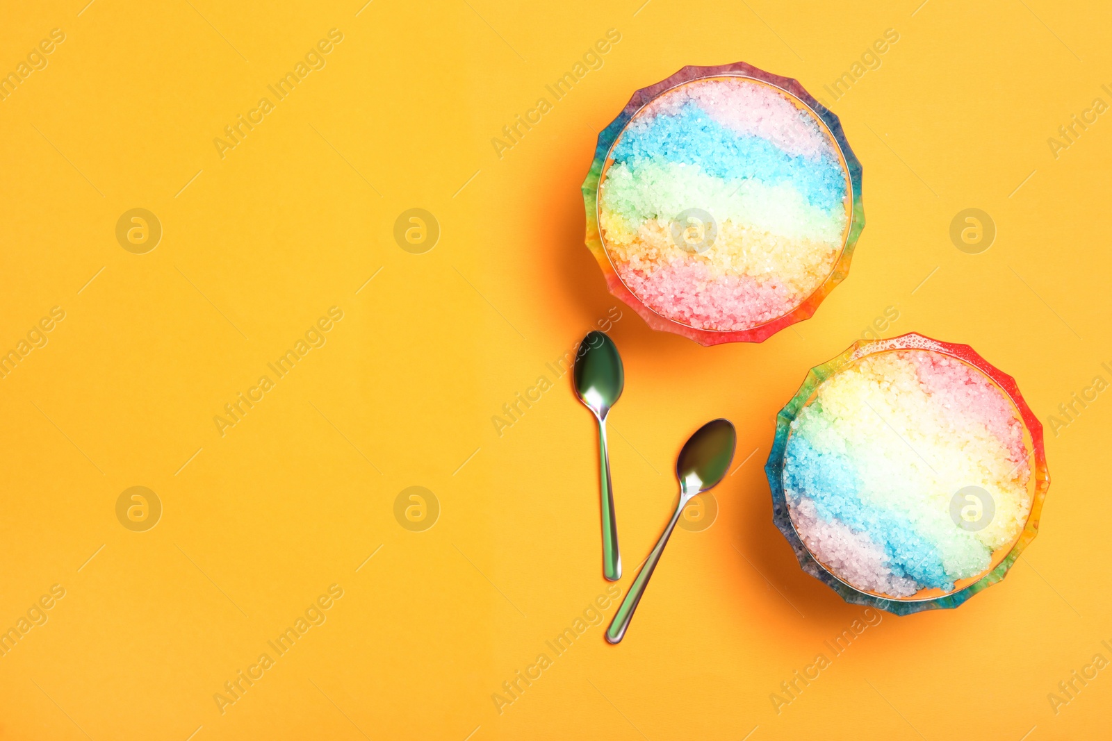 Photo of Rainbow shaving ice in glass dessert bowls and spoons on orange background, flat lay. Space for text