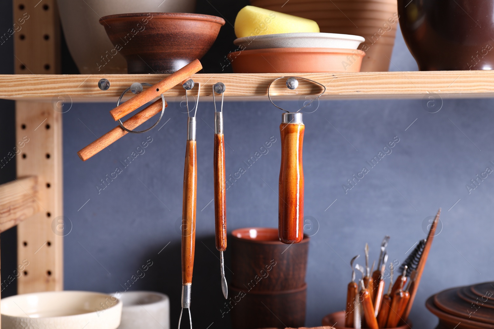 Photo of Set of different crafting tools and clay dishes on wooden rack in workshop, closeup