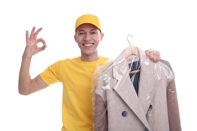 Photo of Dry-cleaning delivery. Happy courier holding coat in plastic bag and showing OK gesture on white background