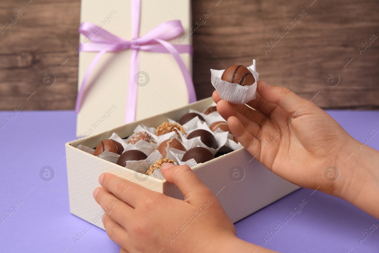 Photo of Child taking delicious chocolate candy from box at light purple table, closeup