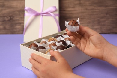 Child taking delicious chocolate candy from box at light purple table, closeup