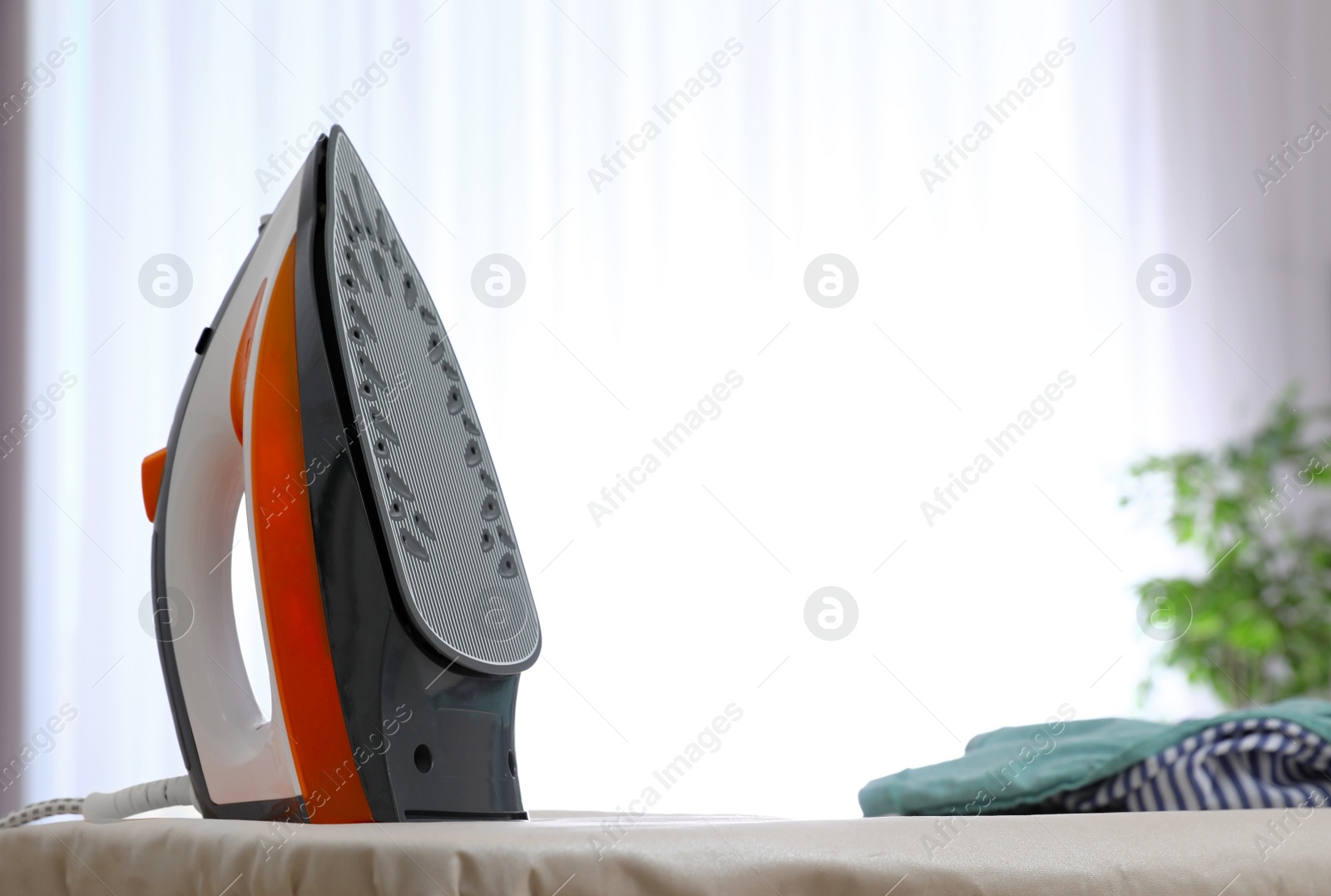 Photo of Modern electric iron and clothes on board indoors. Space for text