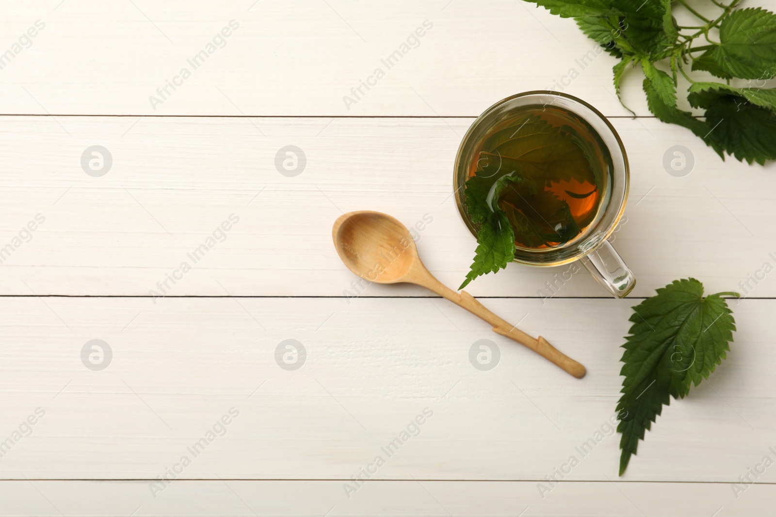 Photo of Glass cup of aromatic nettle tea and green leaves on white wooden table, flat lay. Space for text