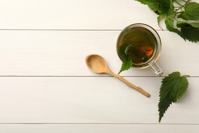 Glass cup of aromatic nettle tea and green leaves on white wooden table, flat lay. Space for text