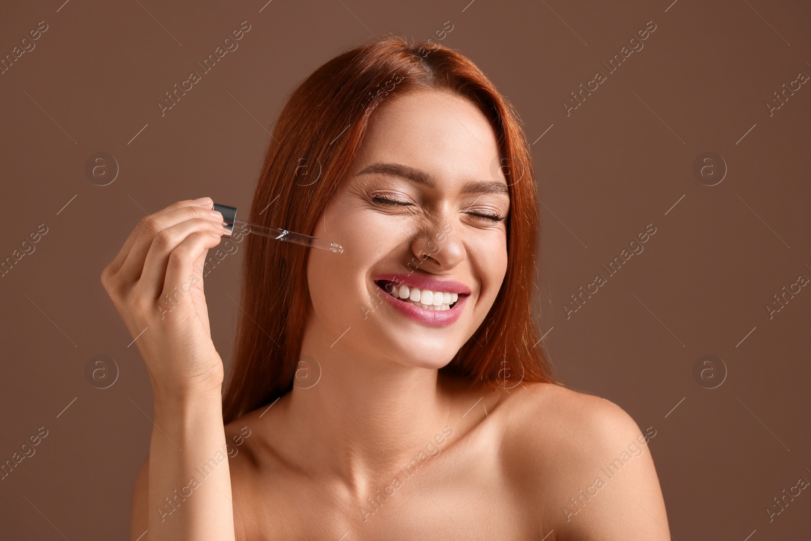 Photo of Beautiful young woman applying cosmetic serum onto her face on brown background