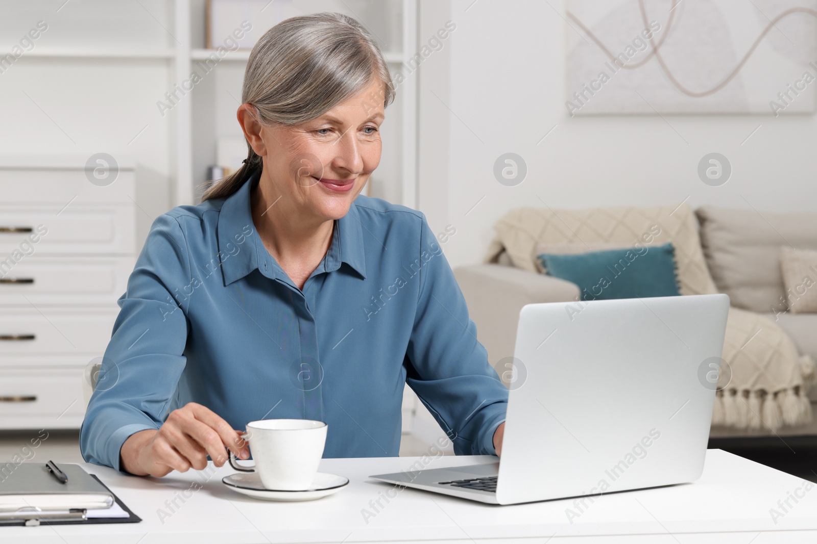 Photo of Beautiful senior woman with cup of drink using laptop at white table indoors