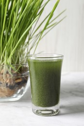 Photo of Wheat grass drink in shot glass and fresh sprouts on white marble table, closeup