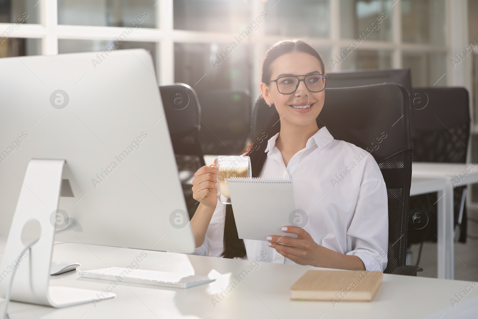 Photo of Happy woman with cup of coffee and notebook near modern computer at white desk in office