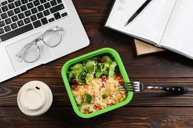 Photo of Container with tasty food, fork, laptop, paper cup of coffee and notebook on wooden table, flat lay. Business lunch