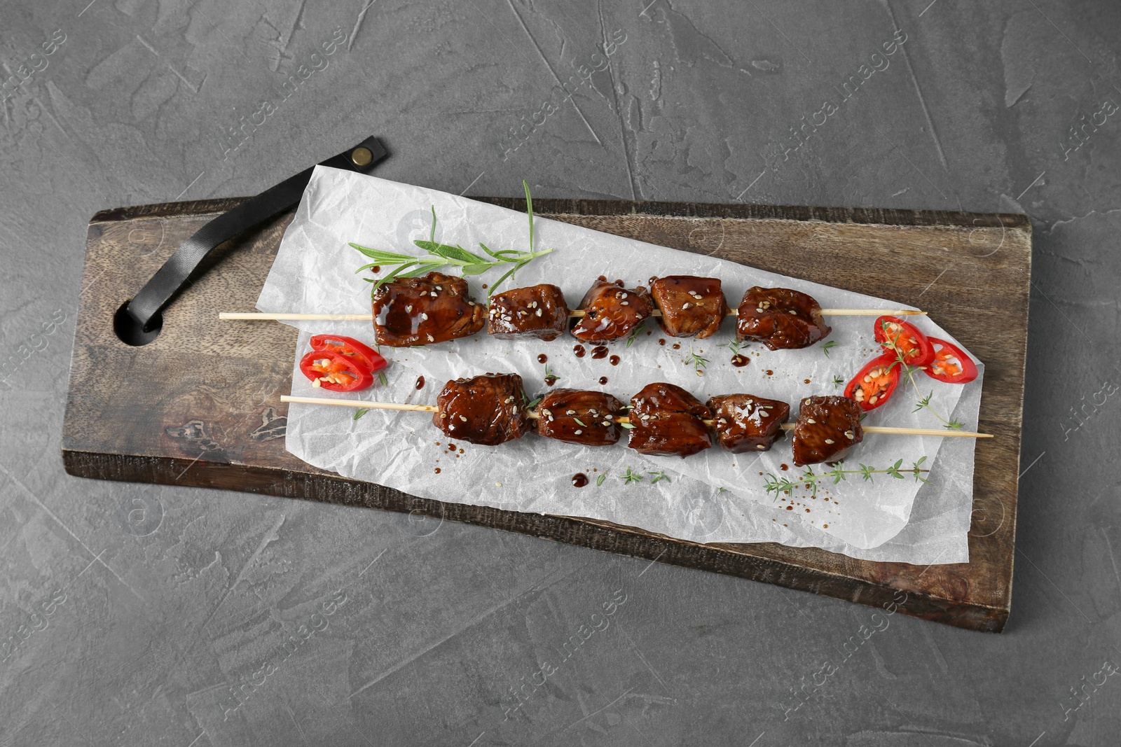 Photo of Skewers with pieces of tasty chicken meat glazed in soy sauce, herbs and cut chili pepper on grey textured table, top view
