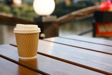 Photo of Takeaway paper cup with coffee on wooden table outdoors. Space for text