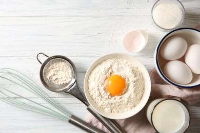 Photo of Flour with yolk in bowl and other ingredients for dough on white wooden table, flat lay
