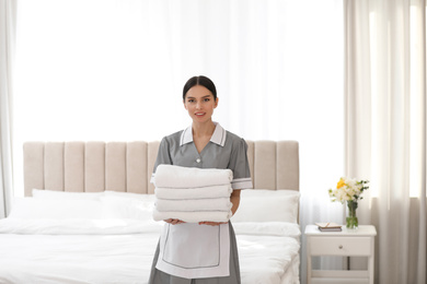 Young chambermaid holding stack of fresh towels in bedroom