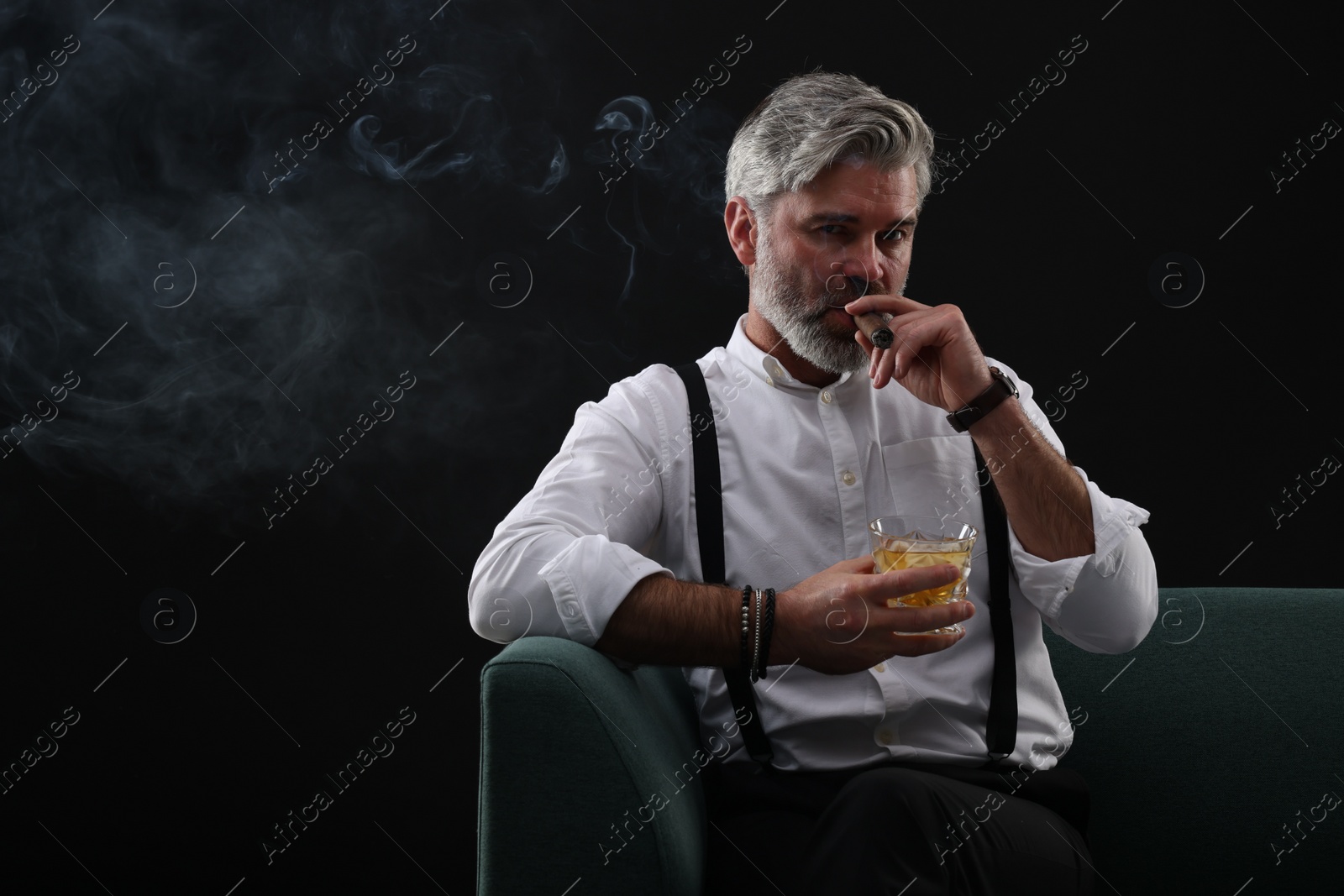 Photo of Bearded man with glass of whiskey smoking cigar on sofa against black background. Space for text