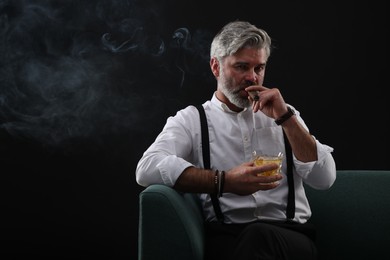 Bearded man with glass of whiskey smoking cigar on sofa against black background. Space for text