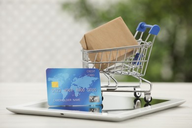 Photo of Online payment concept. Small shopping cart with bank card, box and tablet on white table, closeup