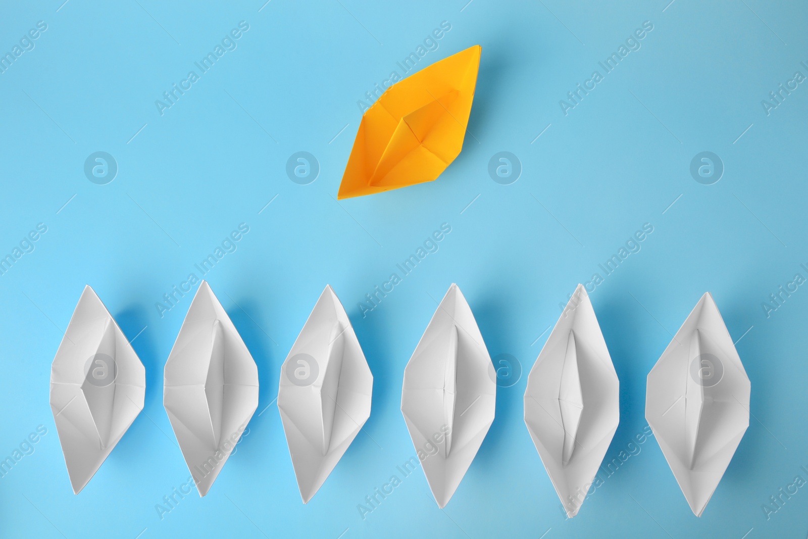 Photo of Yellow paper boat floating away from others on light blue background, flat lay. Uniqueness concept