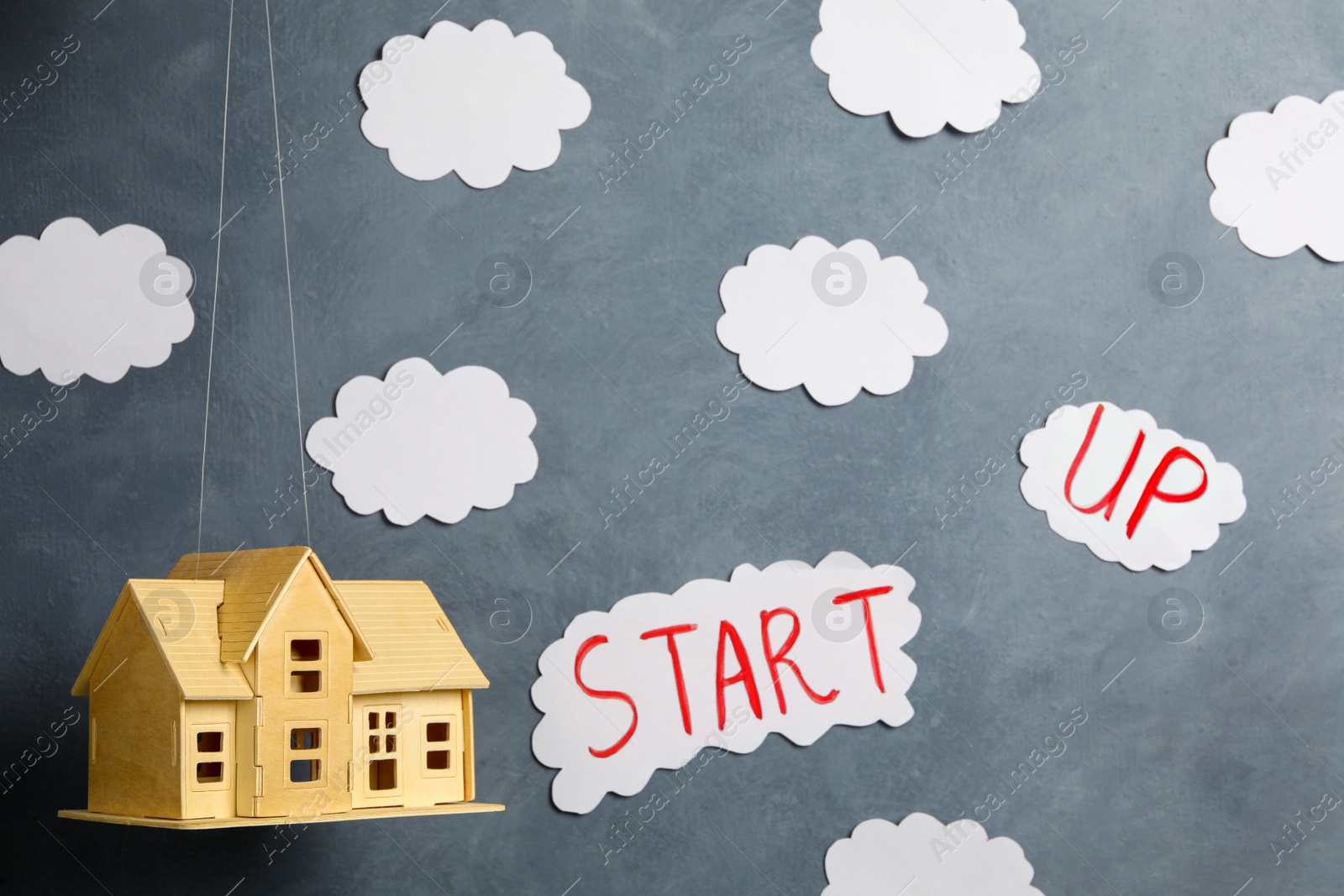 Photo of Wooden house model near grey wall with paper clouds and words Start Up