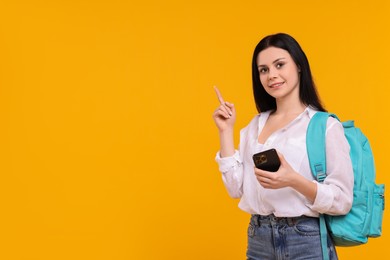 Photo of Smiling student with smartphone pointing at something on yellow background. Space for text