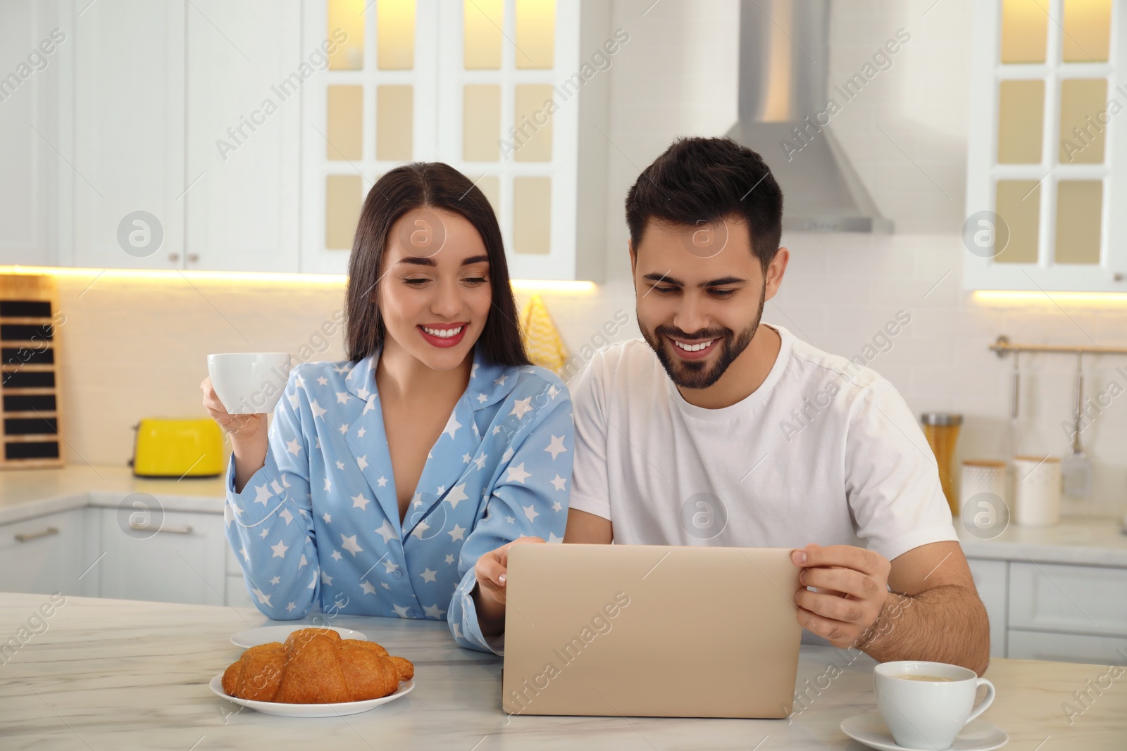 Photo of Happy couple in pajamas with laptop having breakfast at kitchen table