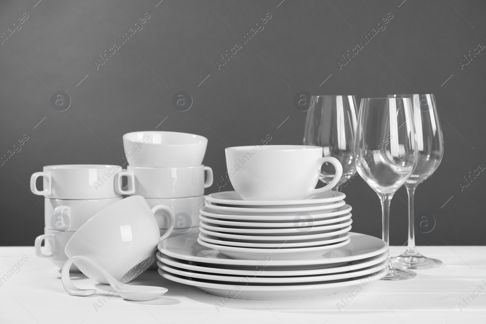 Photo of Set of clean dishware and glasses on white wooden table against grey background