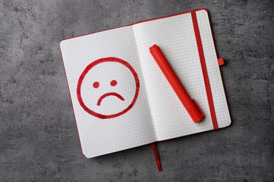 Notebook with drawing of unhappy face and red marker on grey background, top view. Depression symptoms