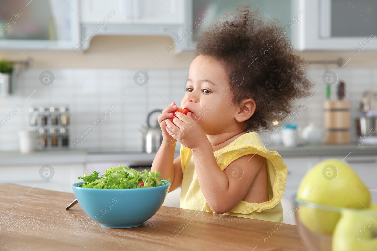 Photo of Cute African-American girl eating vegetable salad at table in kitchen