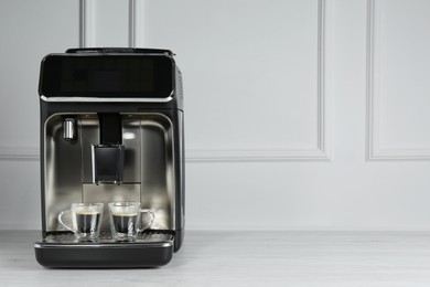 Modern espresso machine with cups of coffee on white wooden table near light wall. Space for text