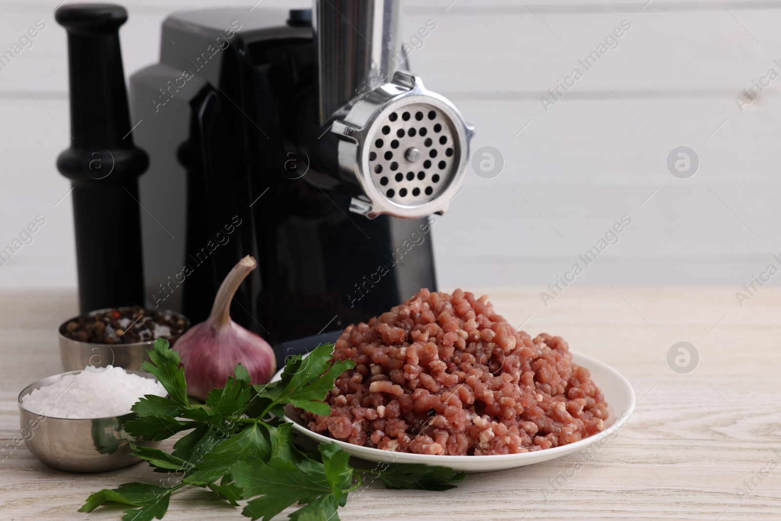 Photo of Electric meat grinder with beef mince, garlic, parsley and spices on white wooden table