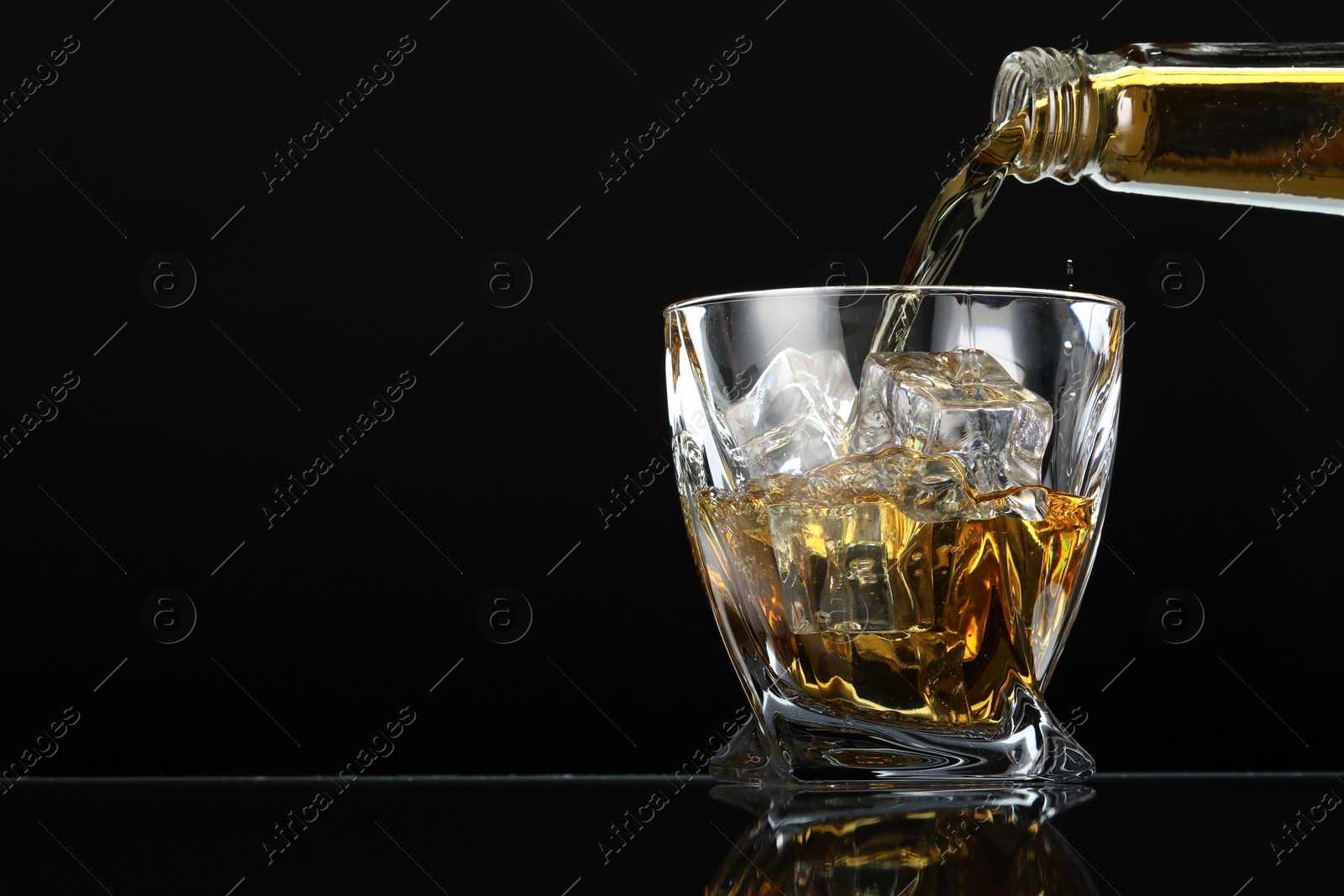 Photo of Pouring whiskey from bottle into glass with ice cubes at table against black background, space for text