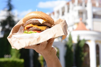 Photo of Woman holding delicious burger in paper wrap outdoors, closeup. Space for text