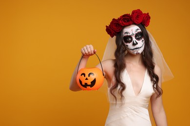 Photo of Young woman in scary bride costume with sugar skull makeup, flower crown and pumpkin bucket on orange background, space for text. Halloween celebration