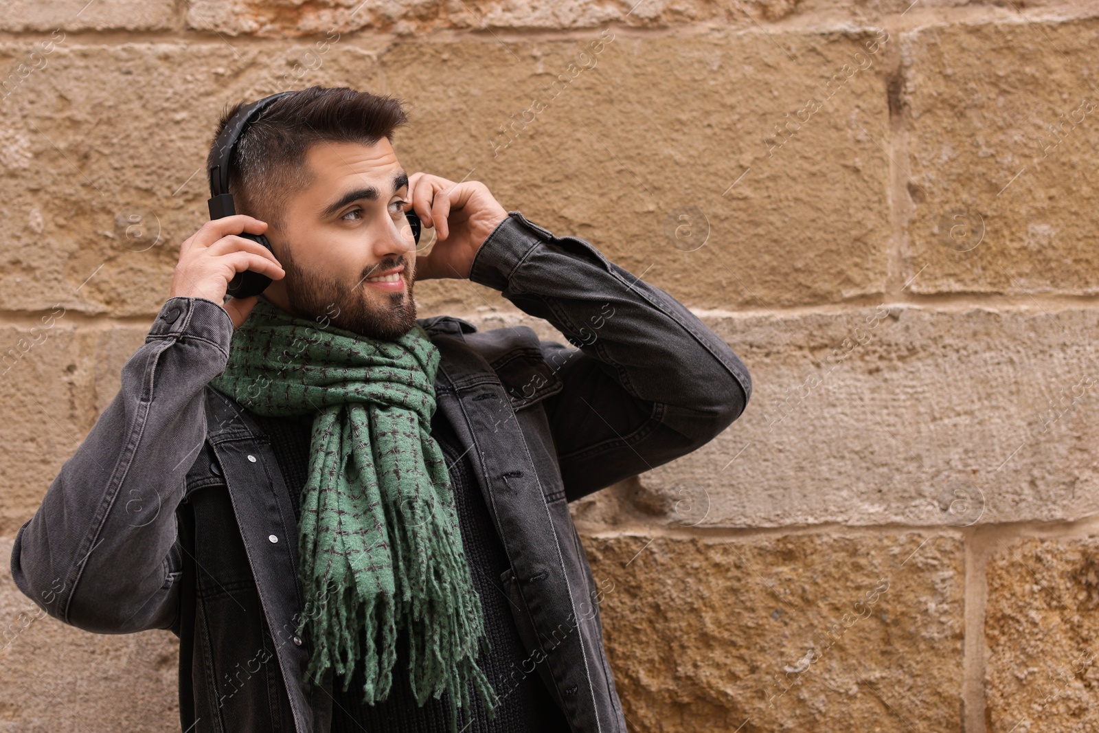 Photo of Smiling man in warm scarf listening to music near brick wall. Space for text